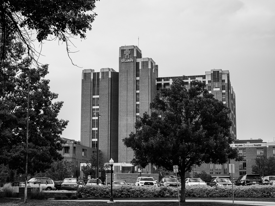 black-and-white photo of hospital building with St. Lukes logo at top, with parking lot and trees