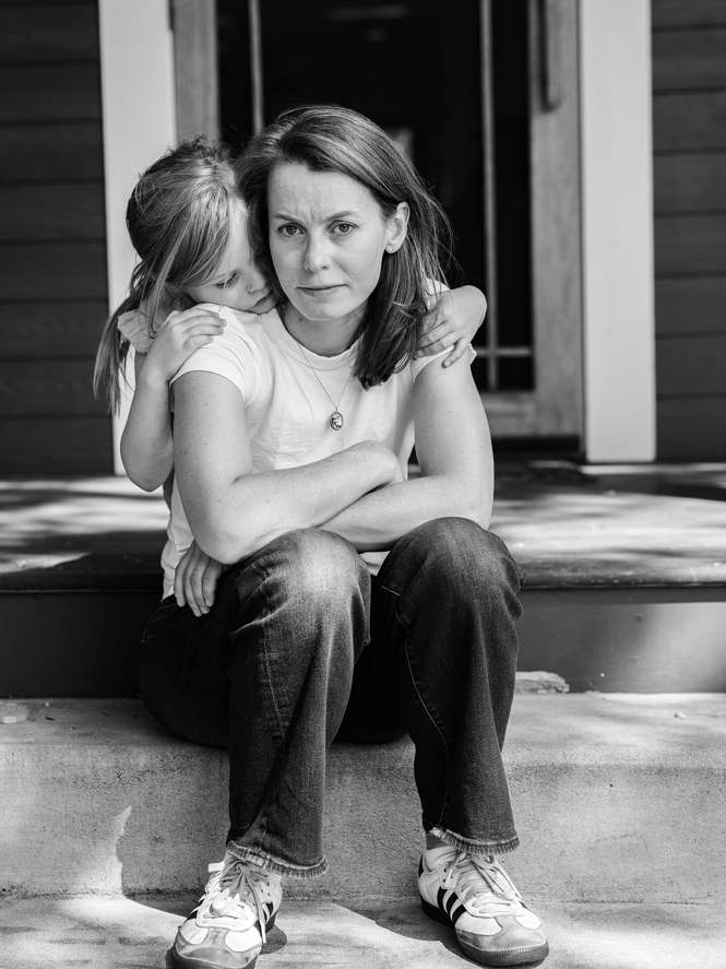 black-and-white photo of woman sitting on steps of front porch with small girl crouching behind her giving her a hug