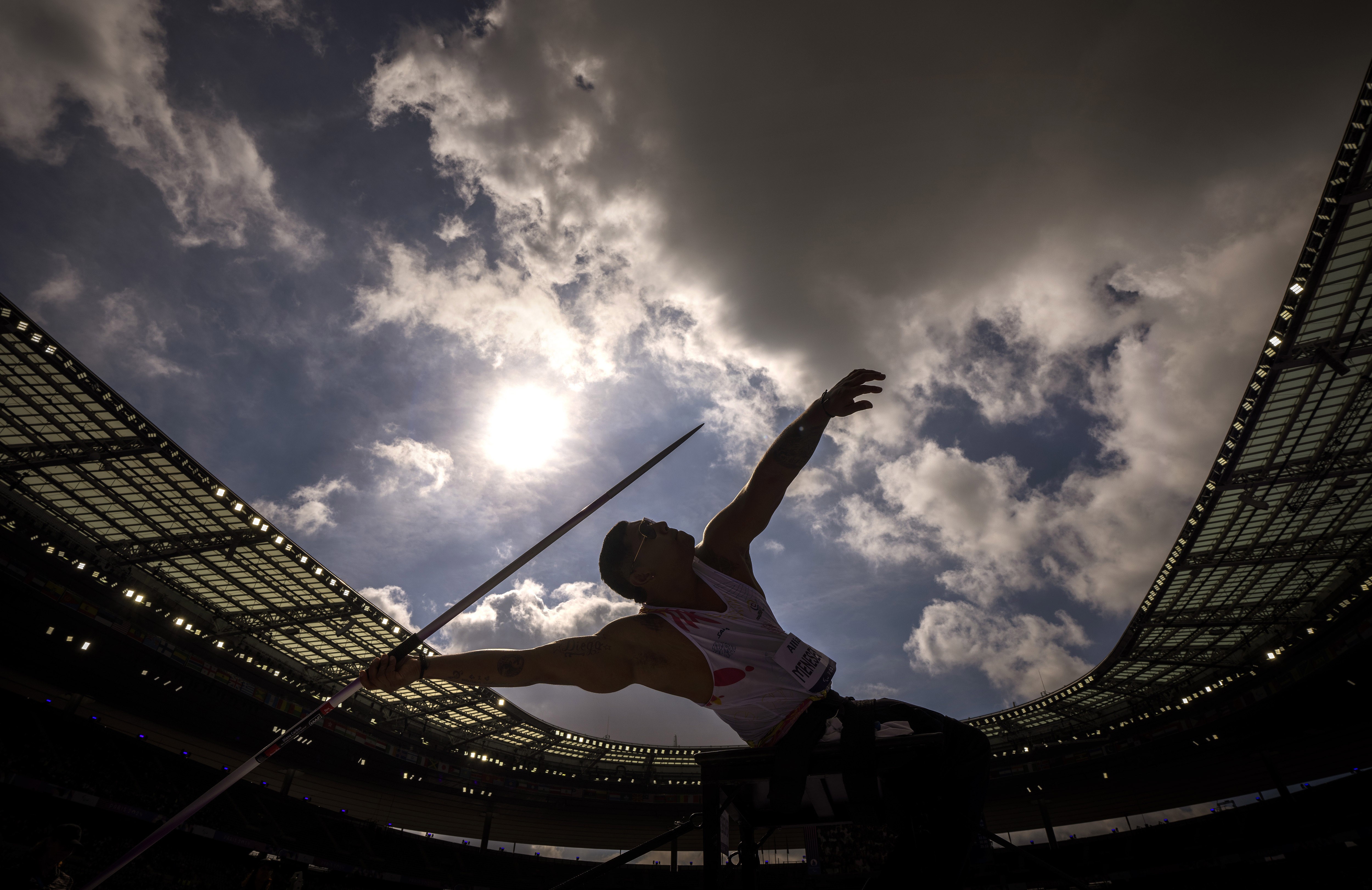 An athlete stretches and leans back while preparing to throw a javelin, seen inside a stadium, silhouetted against a cloudy sky.