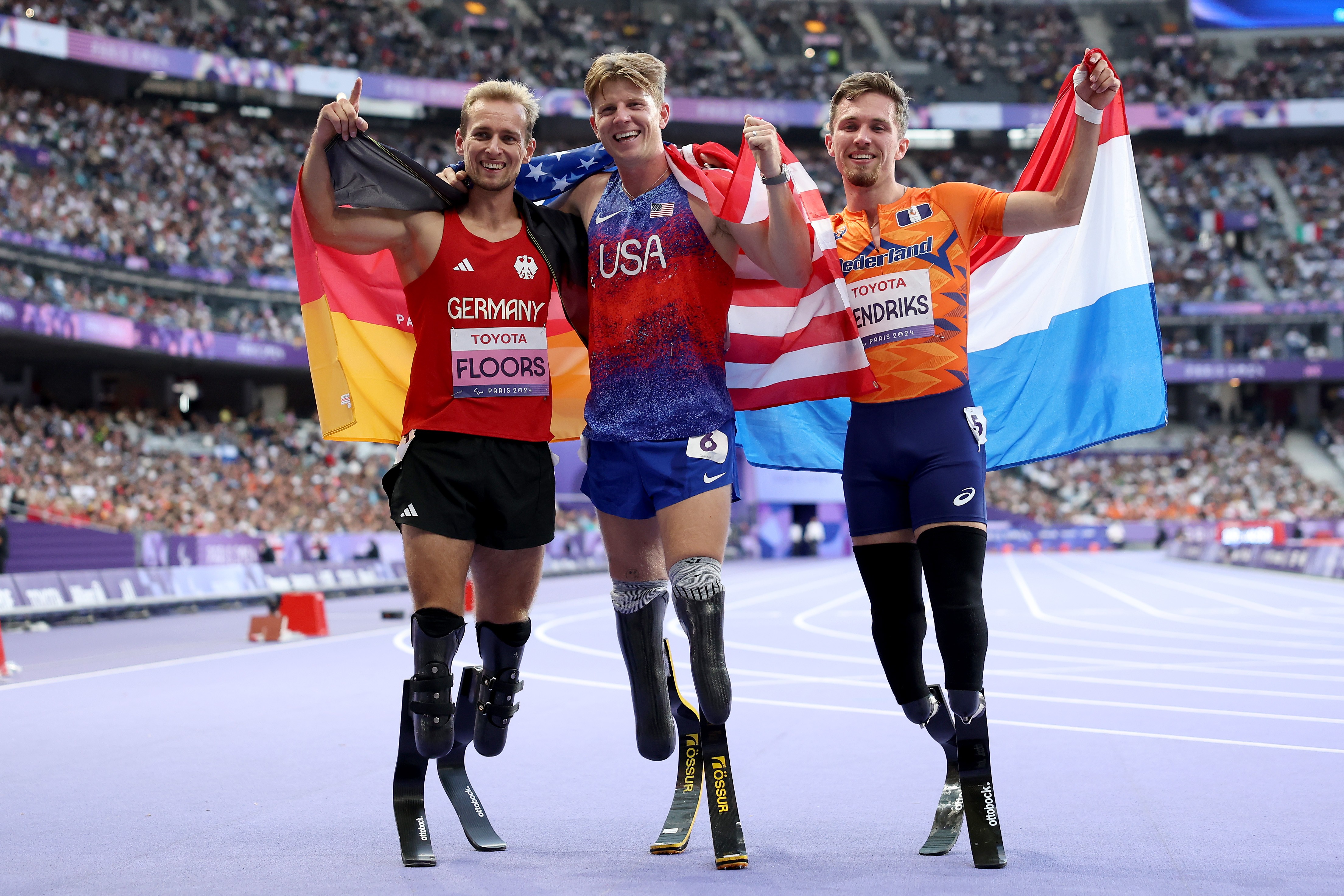 Three para-athletes wearing leg prostheses stand side-by-side, celebrating, draped in their countries' flags, inside a stadium.