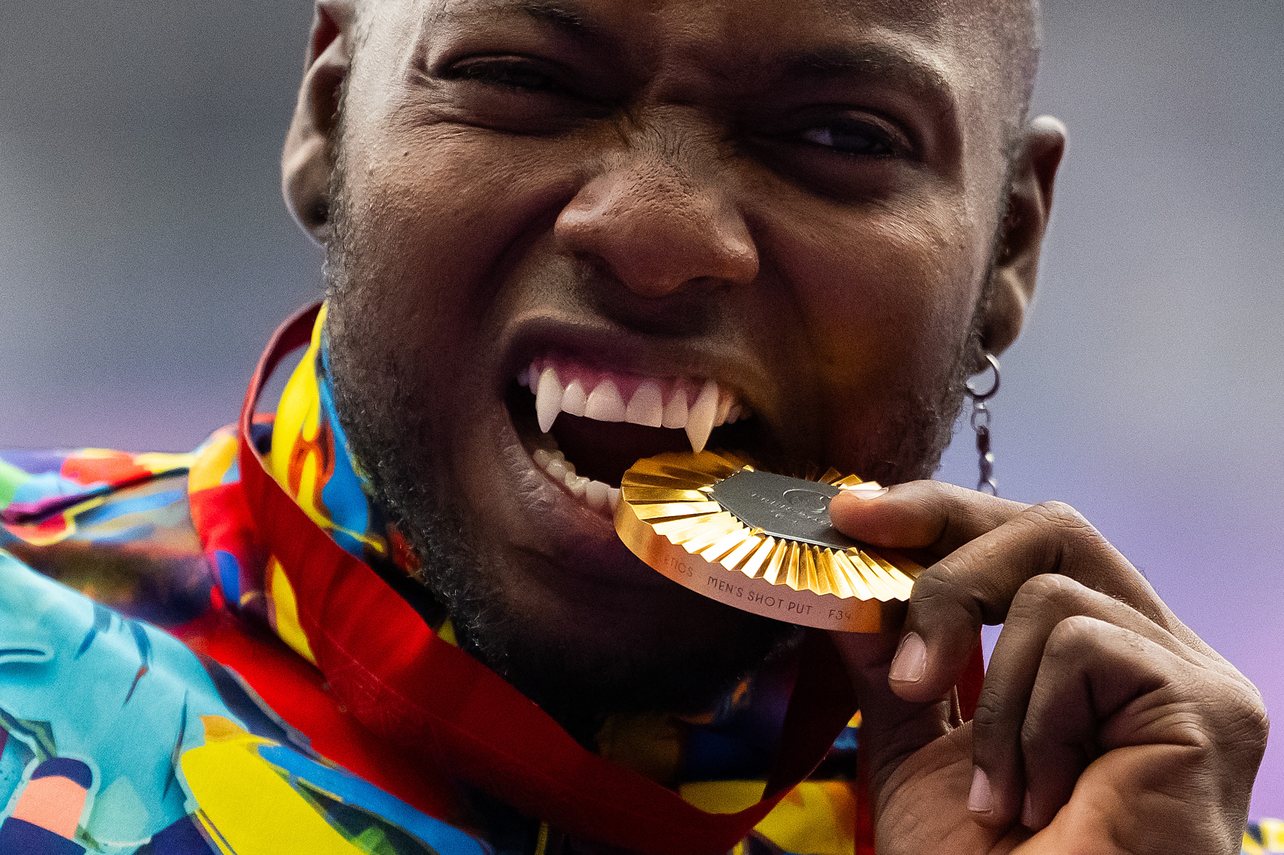 An athlete playfully bites a gold medal during a ceremony. Their canine teeth have been modified to look like long fangs.