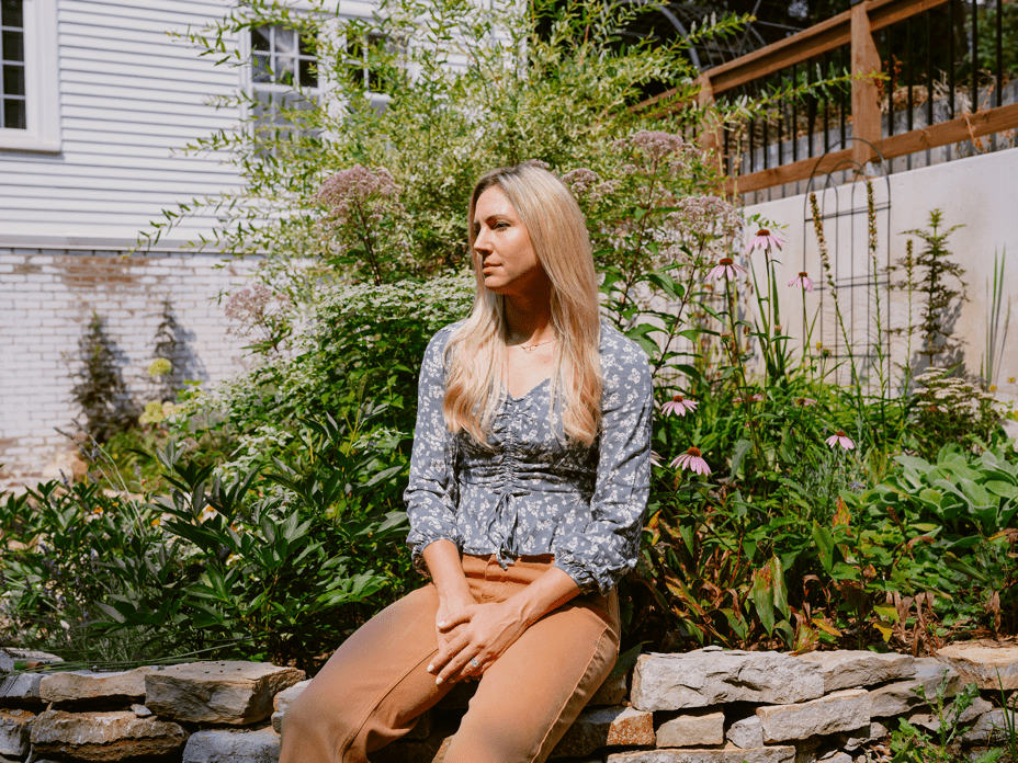 photo of blonde woman sitting outside on stone wall by garden flowers 