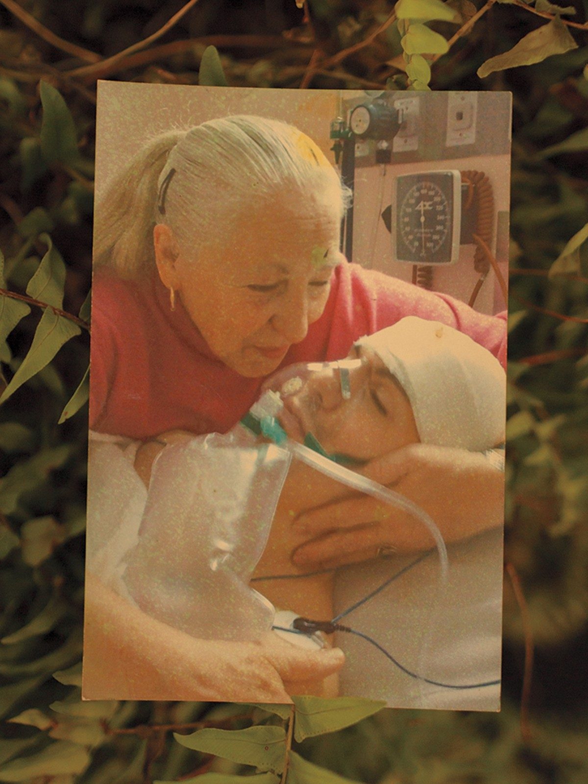 photo of white-haired woman in ponytail cradling bandaged head of man with breathing mask and monitors in her arms