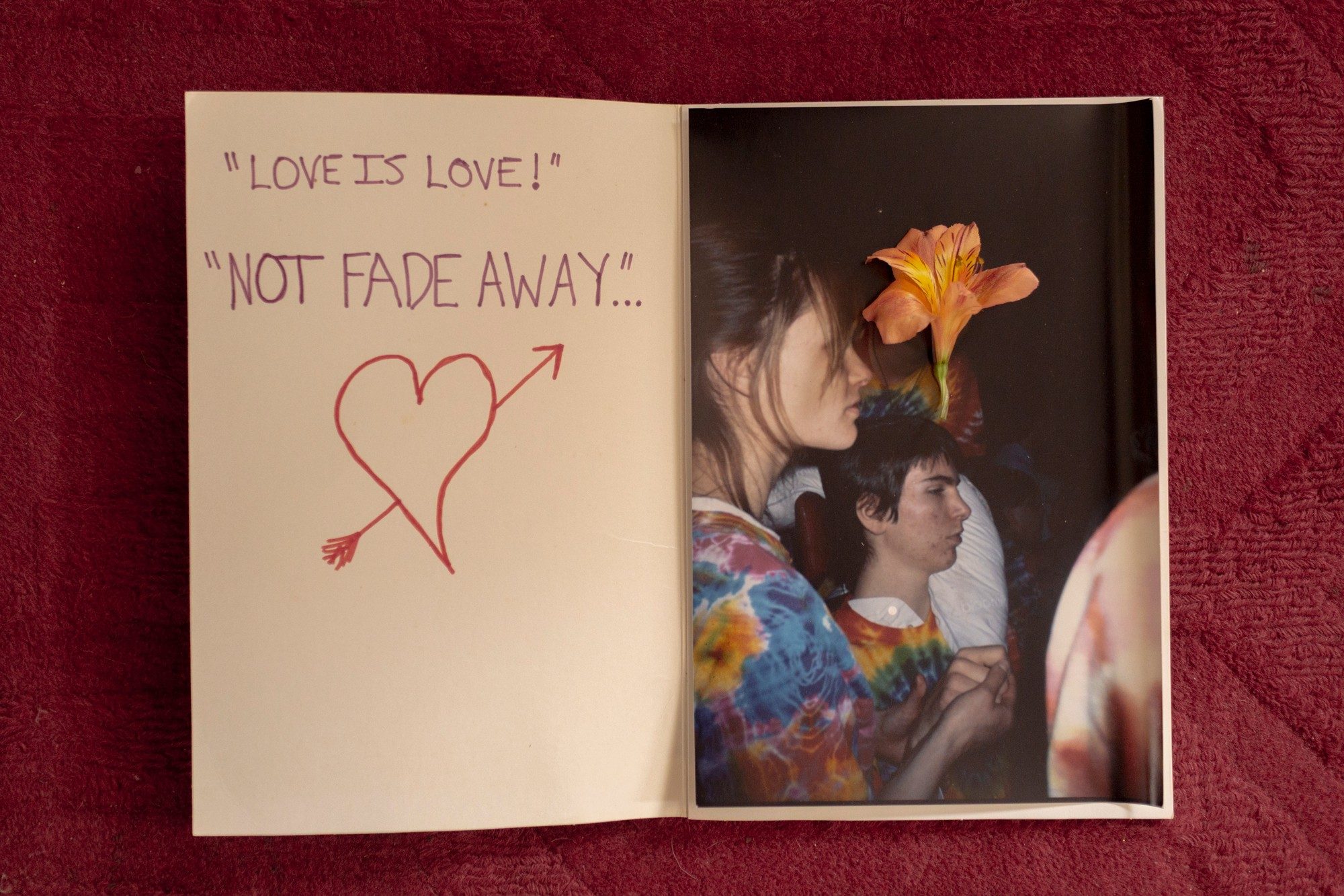 photo of hand-written card with words 'LOVE IS LOVE' AND 'NOT FADE AWAY' alongside heart with arrow on left page and photo of people with flower on right